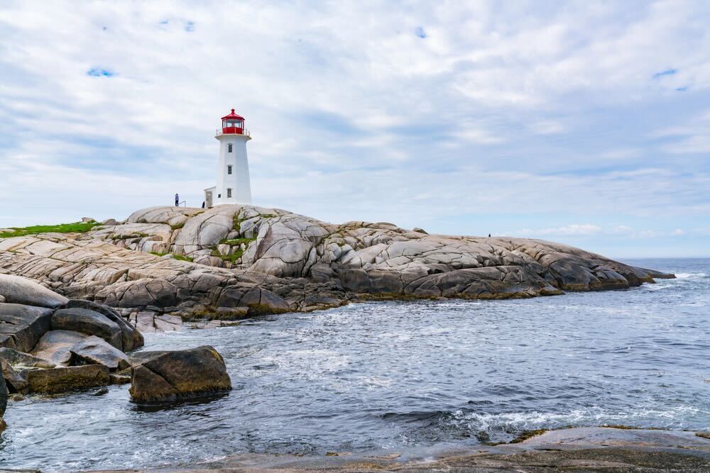 Scenic view of Peggy’s Cove lighthouse and rugged granite shoreline in Nova Scotia, Canada, with waves crashing against the rocks.